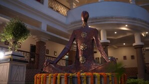 A statue of a seated figure with a flower on the chest, surrounded by a garland, in a building with a curved ceiling and columns. - The Kolam (Bolpur)