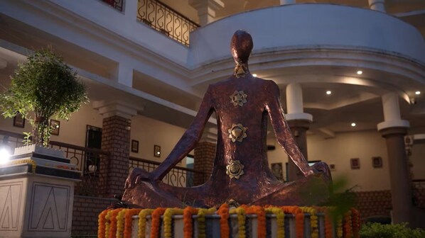 A statue of a seated figure with a flower on the chest, surrounded by a garland, in a building with a curved ceiling and columns. - The Kolam (Bolpur)