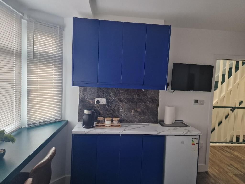 A compact kitchen with blue cabinets, a marble countertop, a coffee maker, and a TV mounted on the wall.