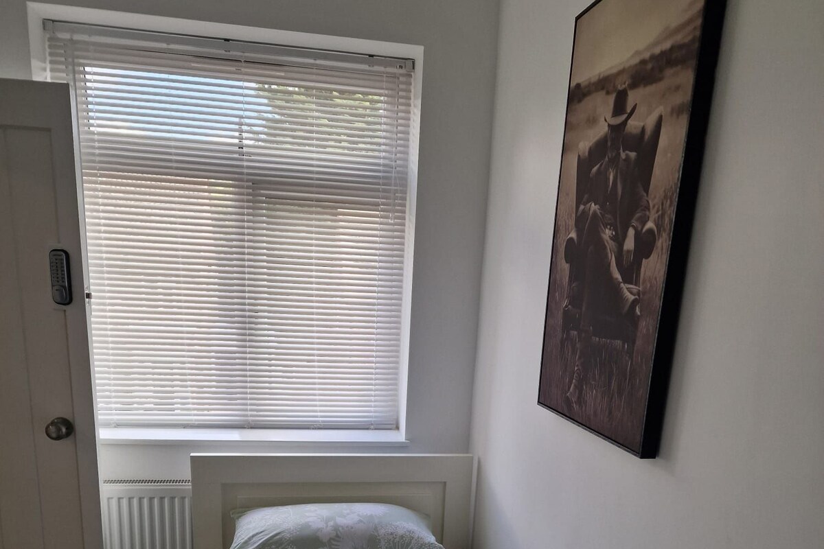 Una cama individual con colcha estampada, una almohada blanca y una toalla doblada sobre la cama. Hay una ventana con persianas parcialmente abiertas y un cuadro en la pared.