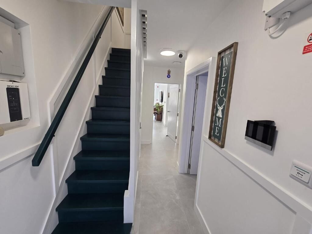 A hallway with a black staircase, a smoke vent, a welcome sign, and a door with a smoke detector.