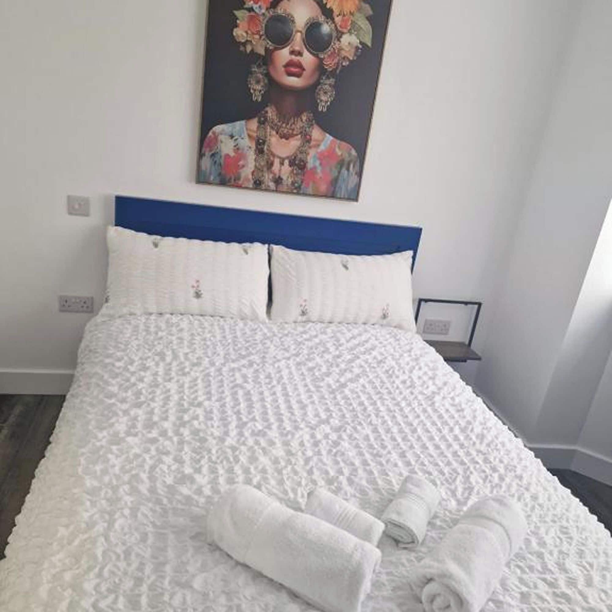 A neatly made bed with white linens and two pillows, a blue headboard, and a framed artwork above it.