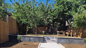 A paved pathway leading to a wooden deck with a table and chairs, surrounded by trees and a wooden fence.