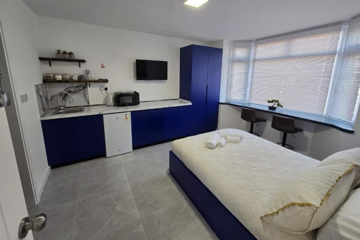 A compact room with a bed, a small kitchenette, a desk, and a window with blinds.
