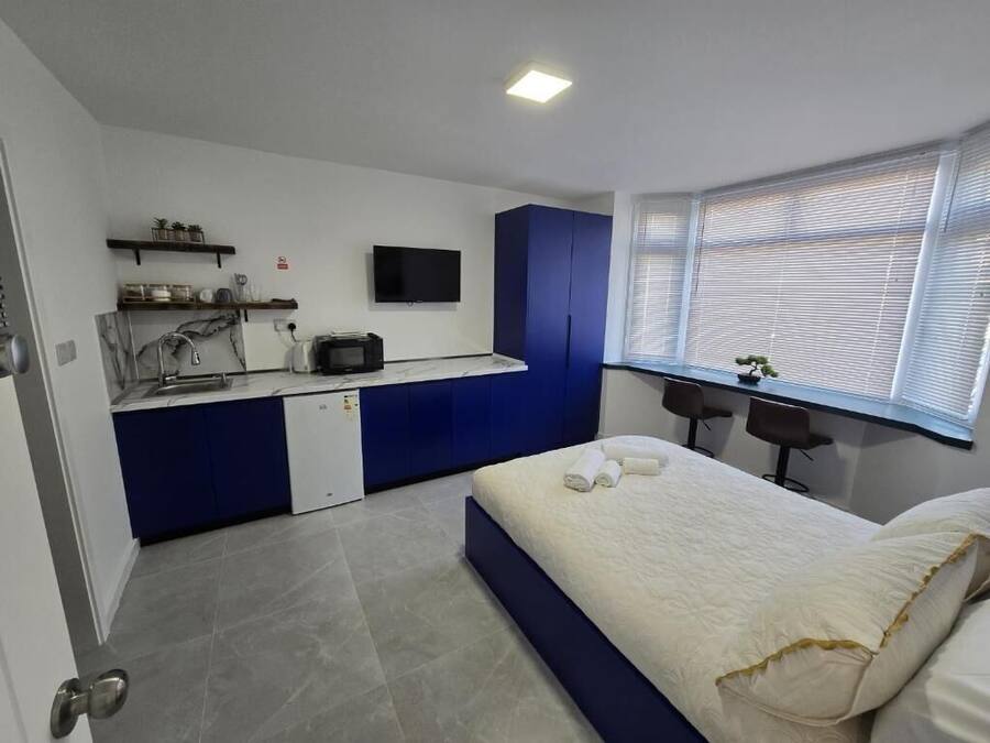 Una habitación compacta con cama, un pequeño espacio kitchenette, un escritorio y una ventana con persianas.