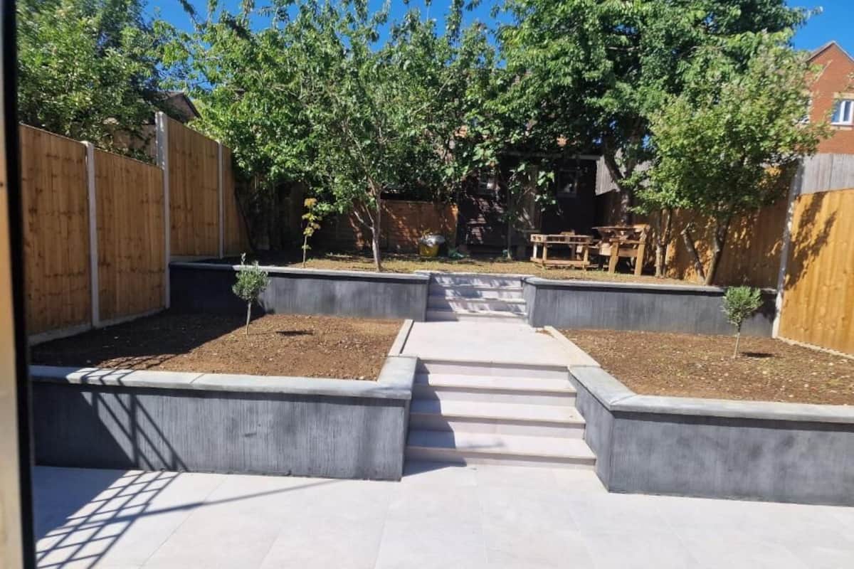 A courtyard with a concrete pathway leading to a wooden table and chairs, surrounded by trees and a wooden fence.