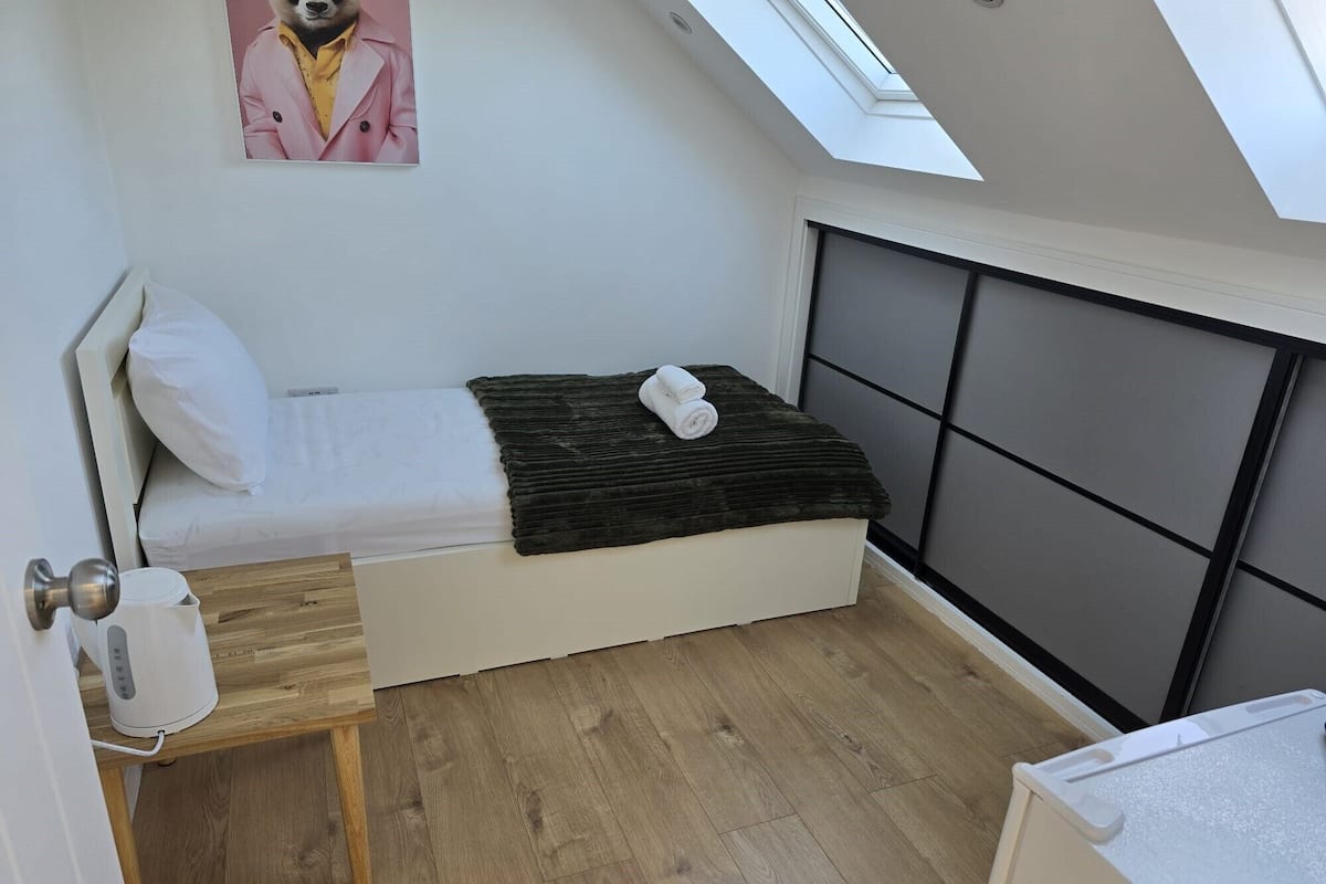 A small, modern bedroom with a skylight, a bed with a dark throw blanket, a bedside table with a white lamp, and a wooden floor.
