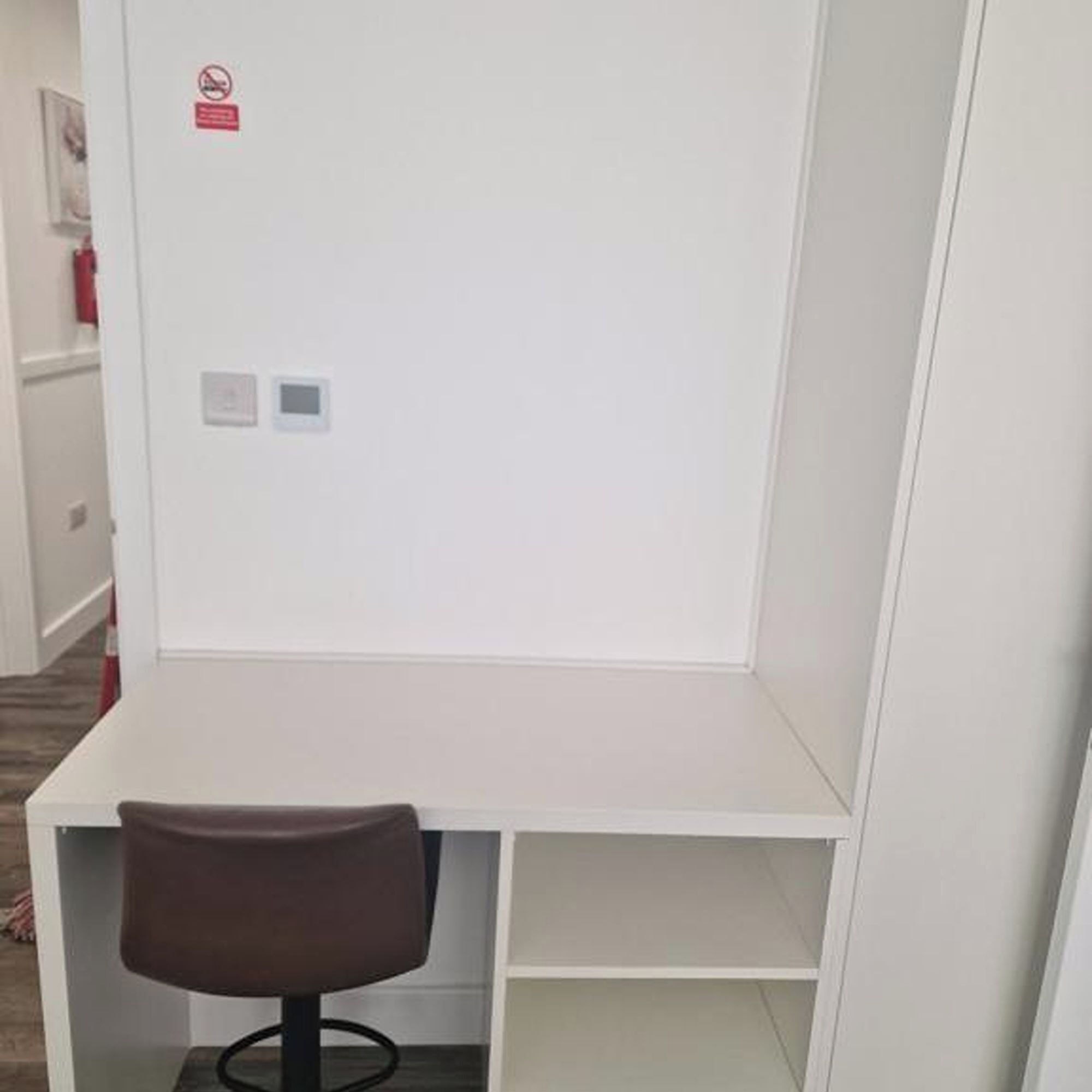 A white desk with a brown chair, a fire alarm, and a small control panel.