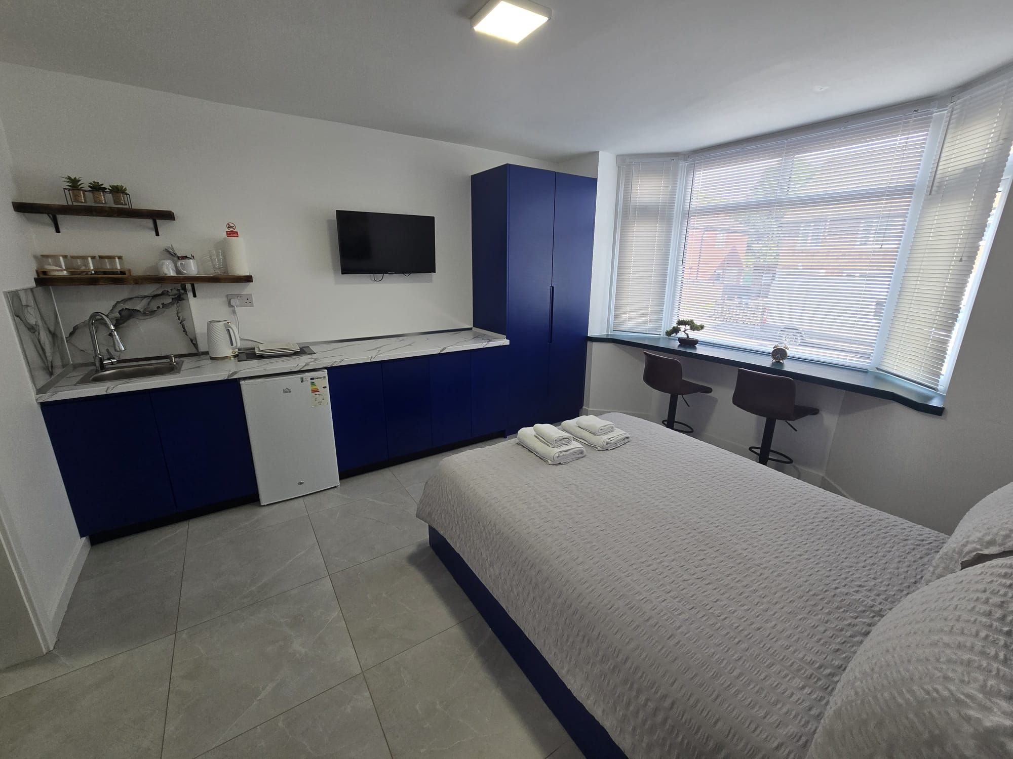 A bedroom with a bed, a desk, a television, and a window with blinds.