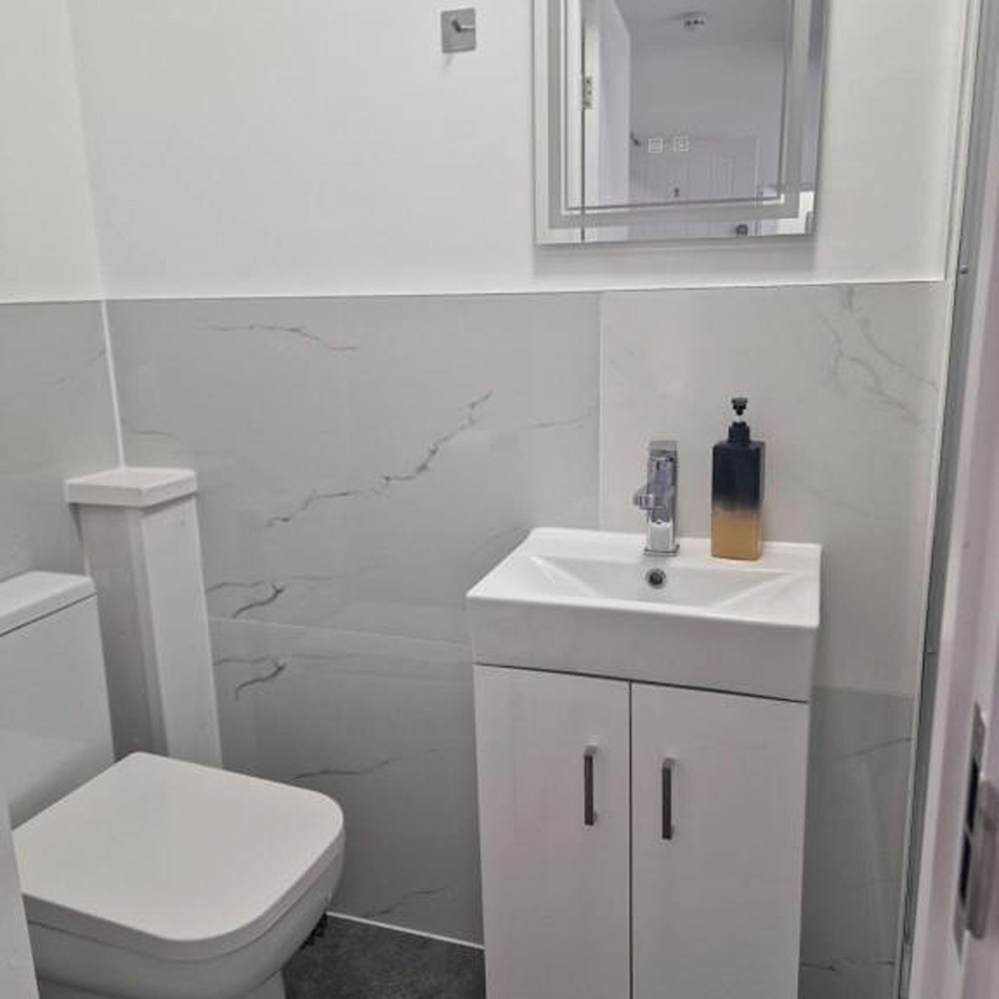 A small, modern bathroom with a white toilet, a white sink with a soap dispenser, and a mirror above the sink.
