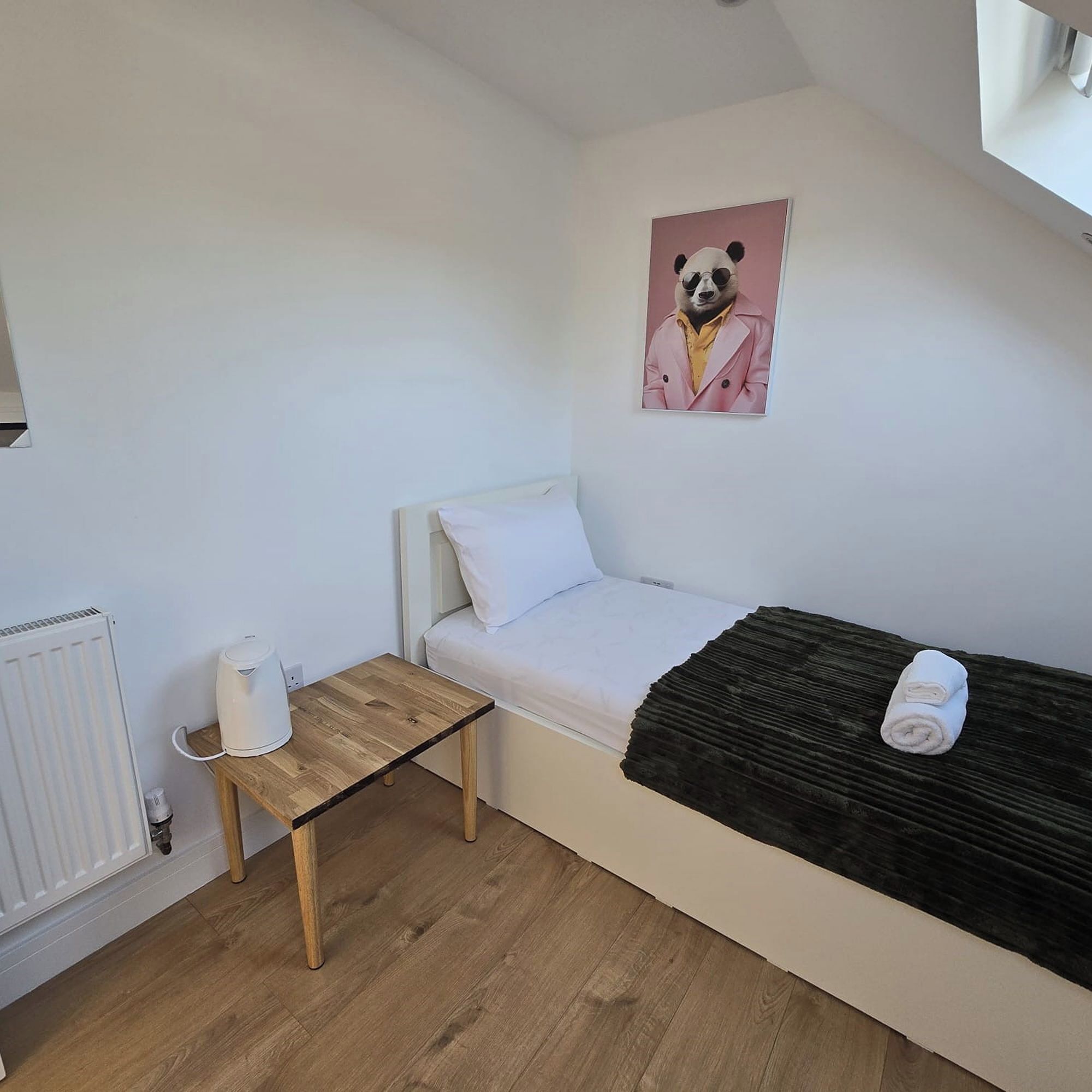 A small, single-bed room with a wooden floor, a white radiator, a small table with a kettle, and a towel on the bed.