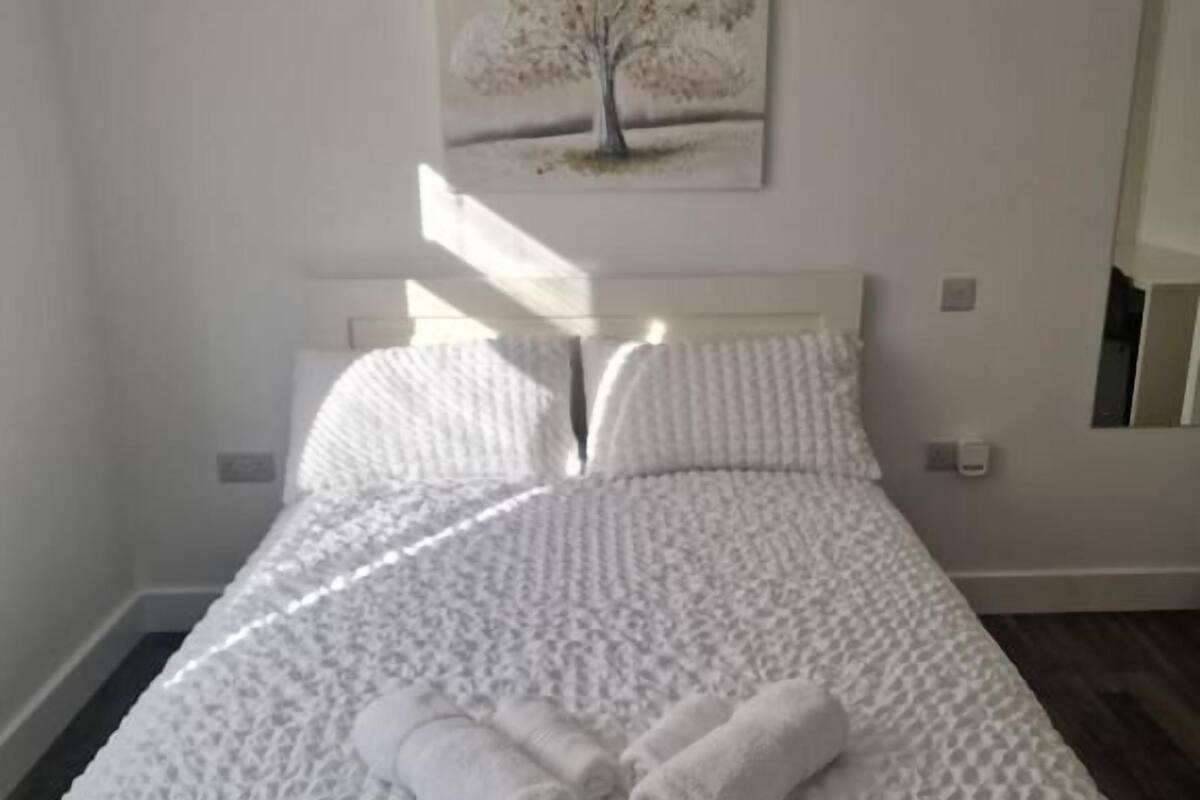 A neatly made bed with a textured white duvet, two rolled towels, and a painting of a tree on the wall above.