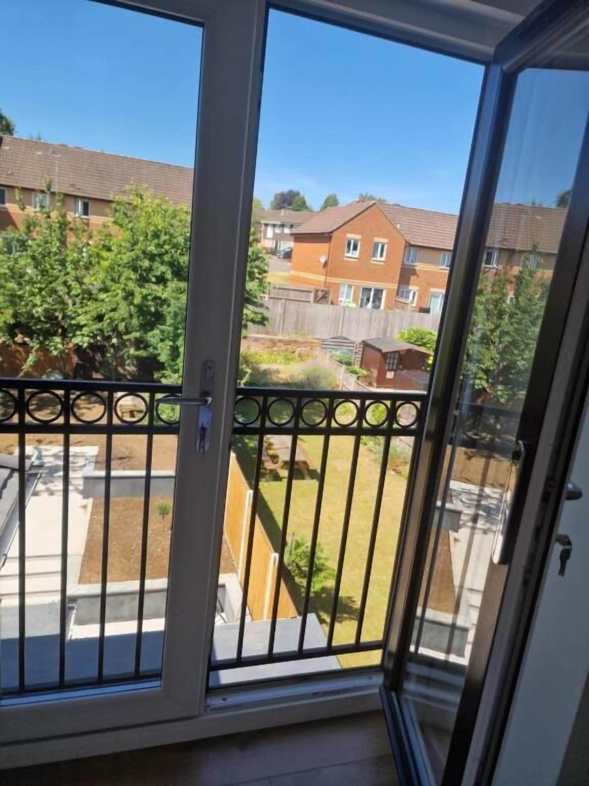 En balkong med ett metallräcke, en skjutdörr i glas och utsikt över ett bostadsområde med hus och träd.