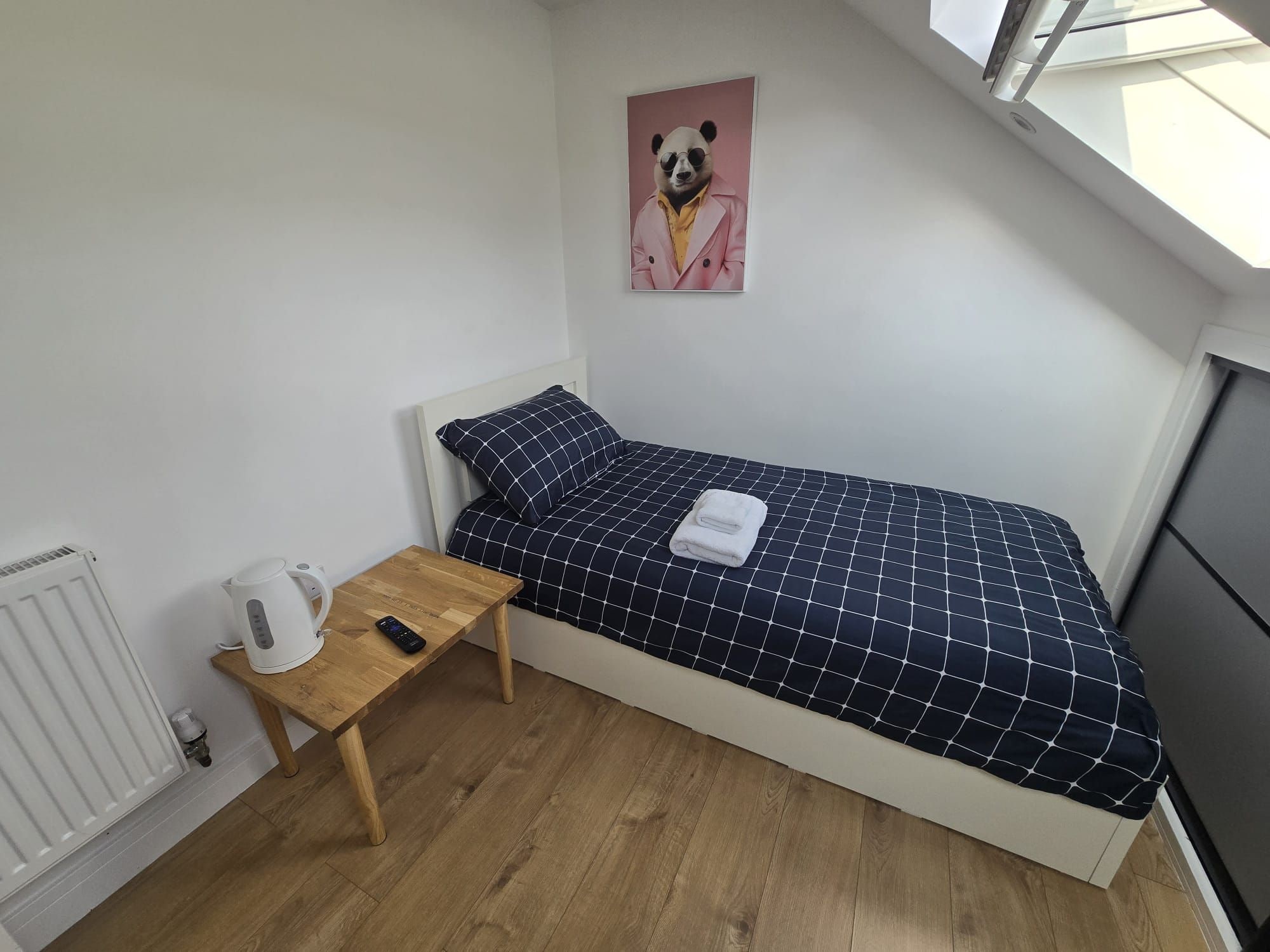 Un dormitorio pequeño y ordenado con una cama individual, una mesita de noche con un hervidor, un control remoto y un radiador.