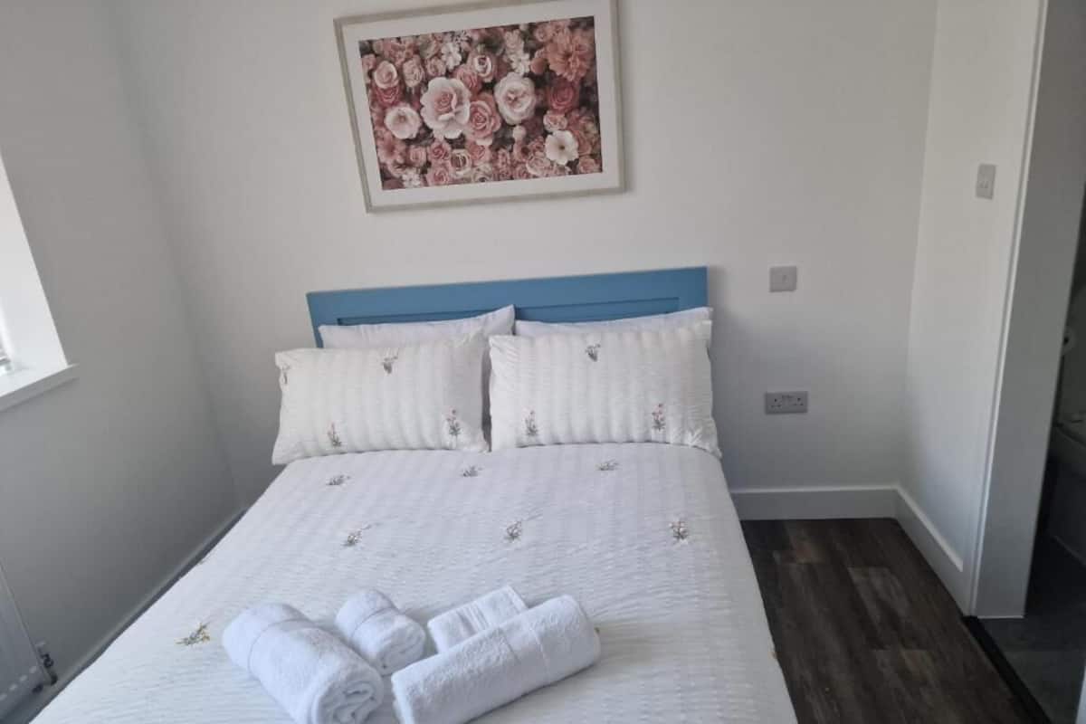 A neatly made bed with white bedding and a blue headboard, a framed floral artwork above it, and a pair of rolled towels on the bed.