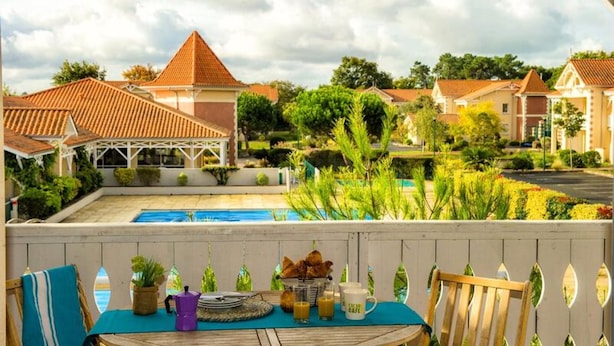 A poolside view with a table set for breakfast, a pergola, and houses with red-tiled roofs in the background. - Apartment in Soulac-sur-mer Near Beaches (Soulac-sur-Mer)