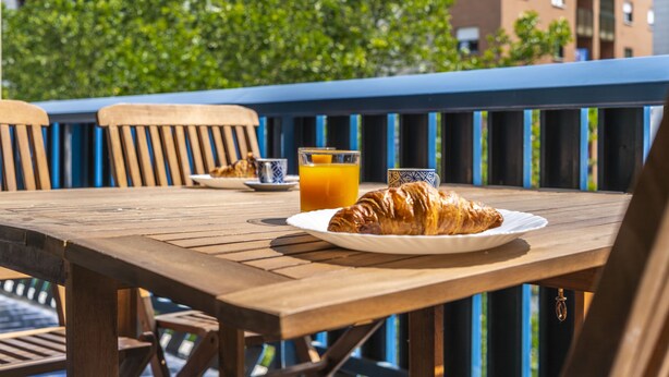 Un tavolo di legno con un piatto di croissant e un bicchiere di succo d'arancia, su un balcone esterno con ringhiere blu e vista su edifici e alberi.