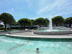 Una fontana con getti d'acqua in una vasca circolare, circondata da alberi e un edificio sullo sfondo.