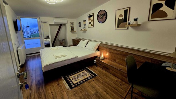 A bedroom with a wooden floor, a bed with white linens, a bedside table with a lamp, a chair, and a TV displaying a scenic view.