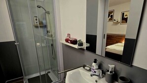 A modern bathroom with a glass shower enclosure, a white sink with a mirror above, and a small shelf with toiletries.