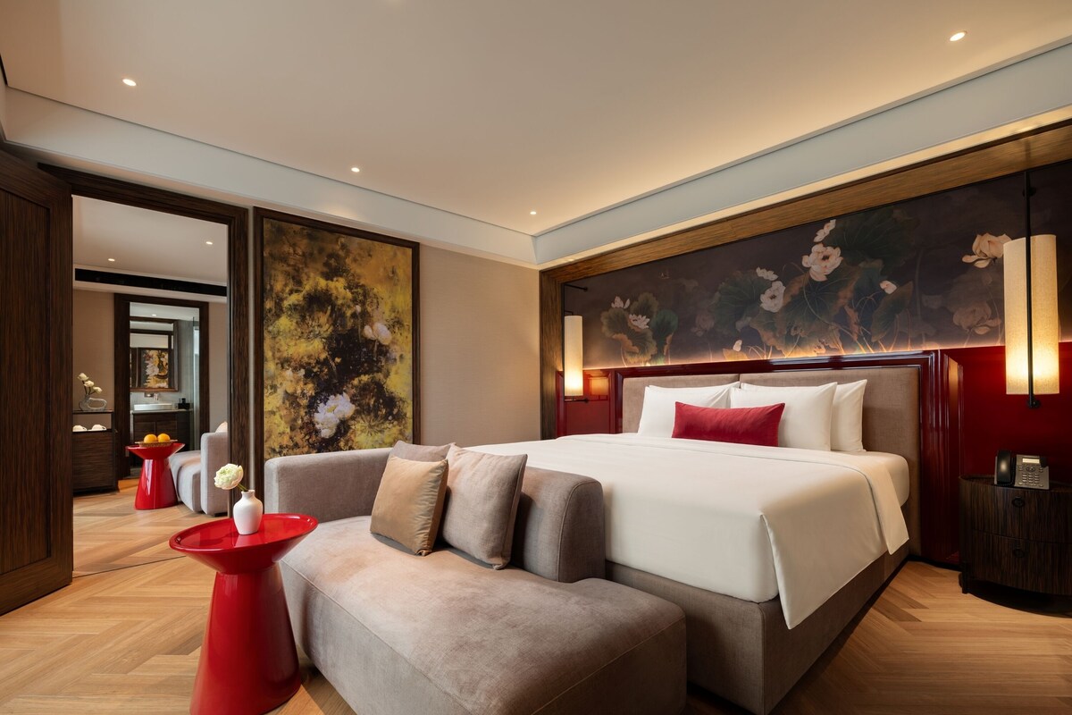 Una habitación de hotel moderna con una cama grande, un sofá, una mesita auxiliar roja y un cuadro en la pared.
