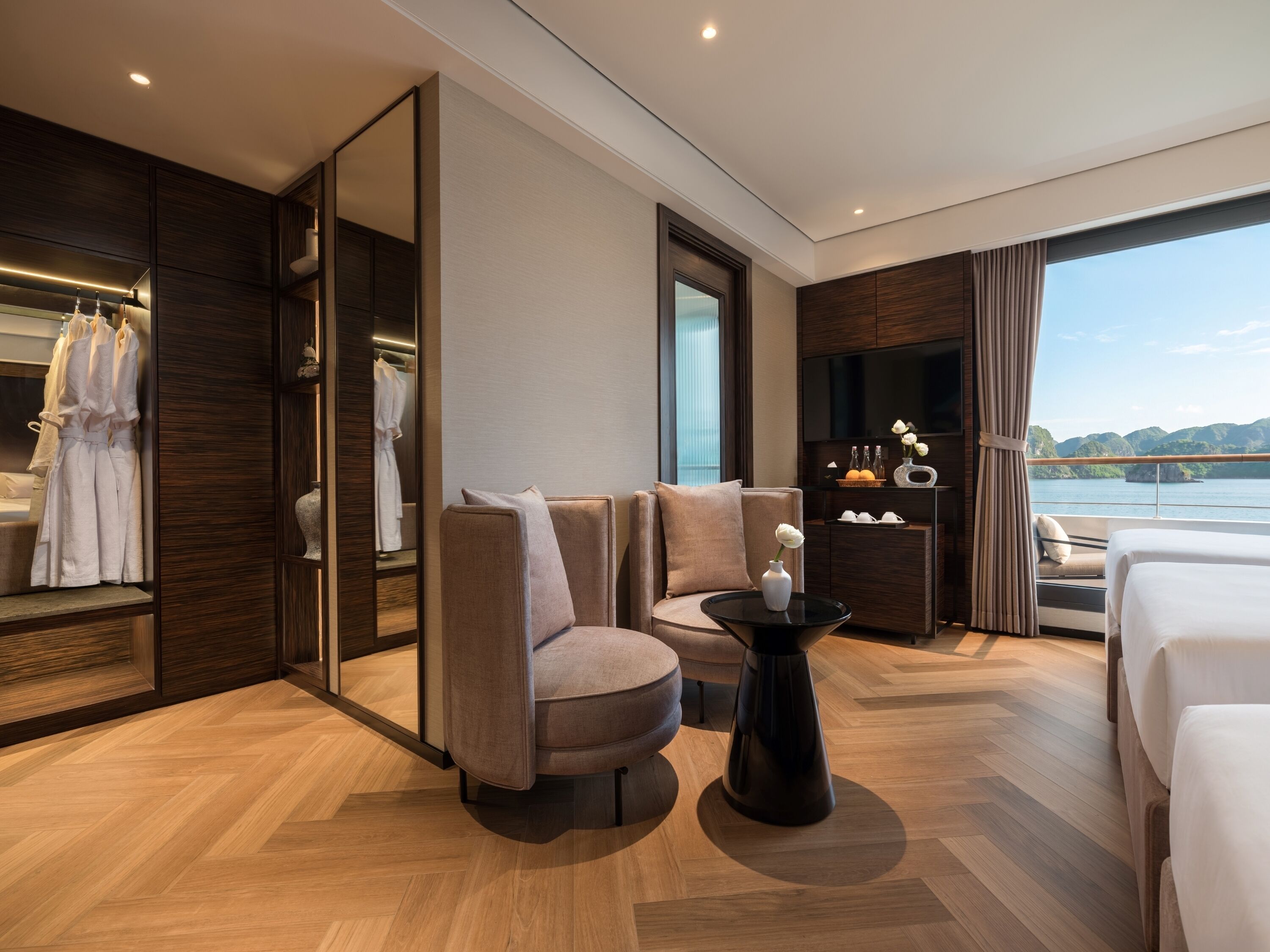 Una habitación de hotel moderna con una cama grande, un espacio de descanso con sofá y sillones, una mesita con un florero, un televisor y vista al agua.