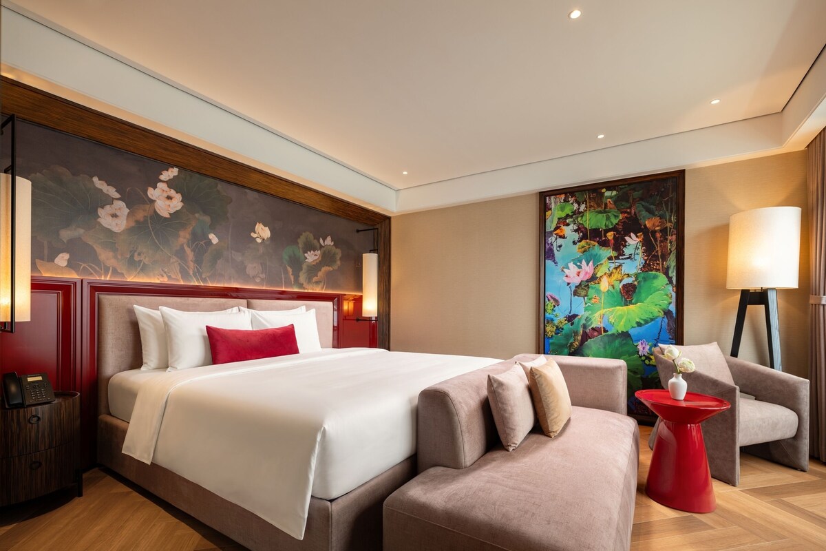 Habitación de hotel con una cama grande, un sofá, una mesita de noche roja y un cuadro en la pared.