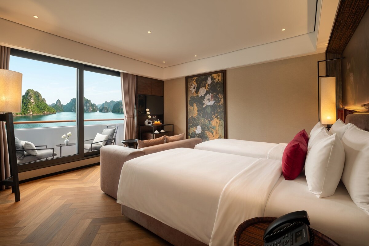 Habitación de hotel con un ventanal que ofrece vistas a un cuerpo de agua y montañas, una cama con ropa blanca y almohadas rojas, una mesita con un florero y un cuadro en la pared.