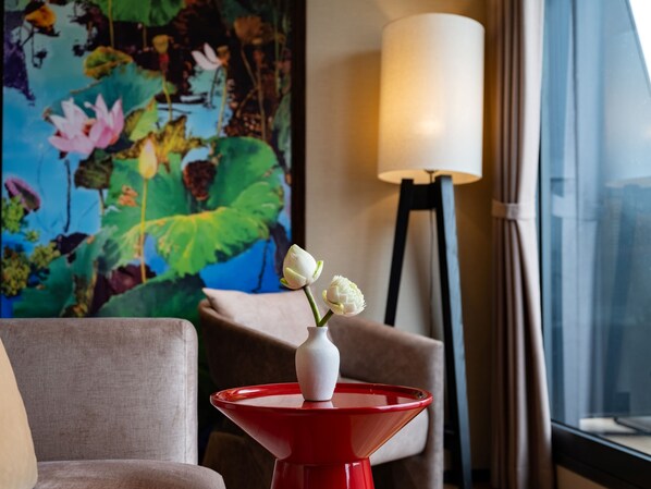 Una habitación con una lámpara de mesa roja, un jarrón con flores, un cuadro y vistas a través de una ventana.