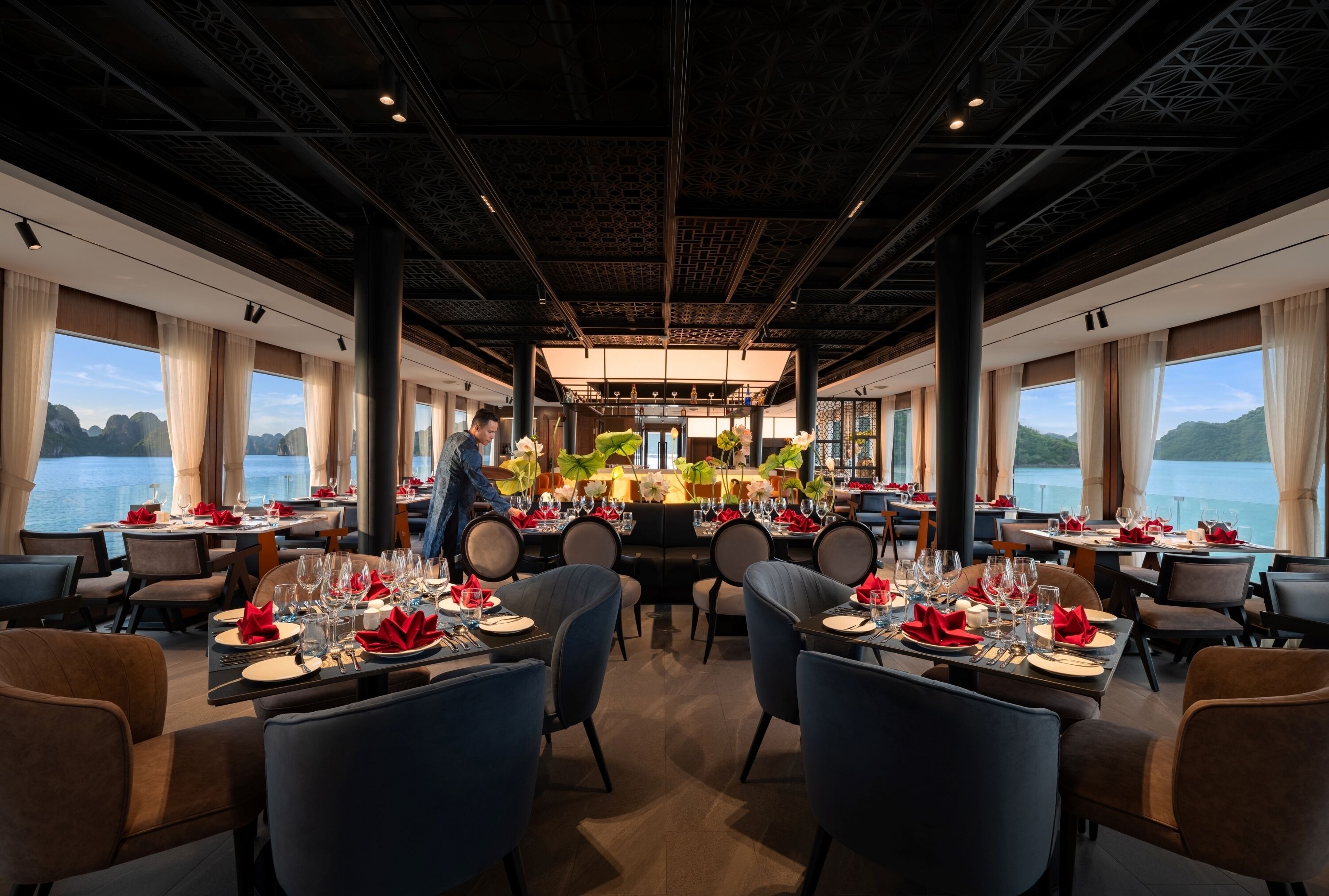Restaurante com mesas preparadas para refeições, grandes janelas com vista para a água e teto alto com padrão geométrico.