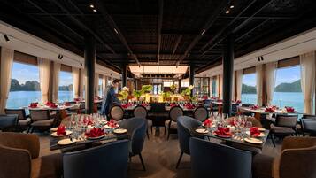 Restaurante com mesas preparadas para refeições, grandes janelas com vista para a água e teto alto com padrão geométrico.