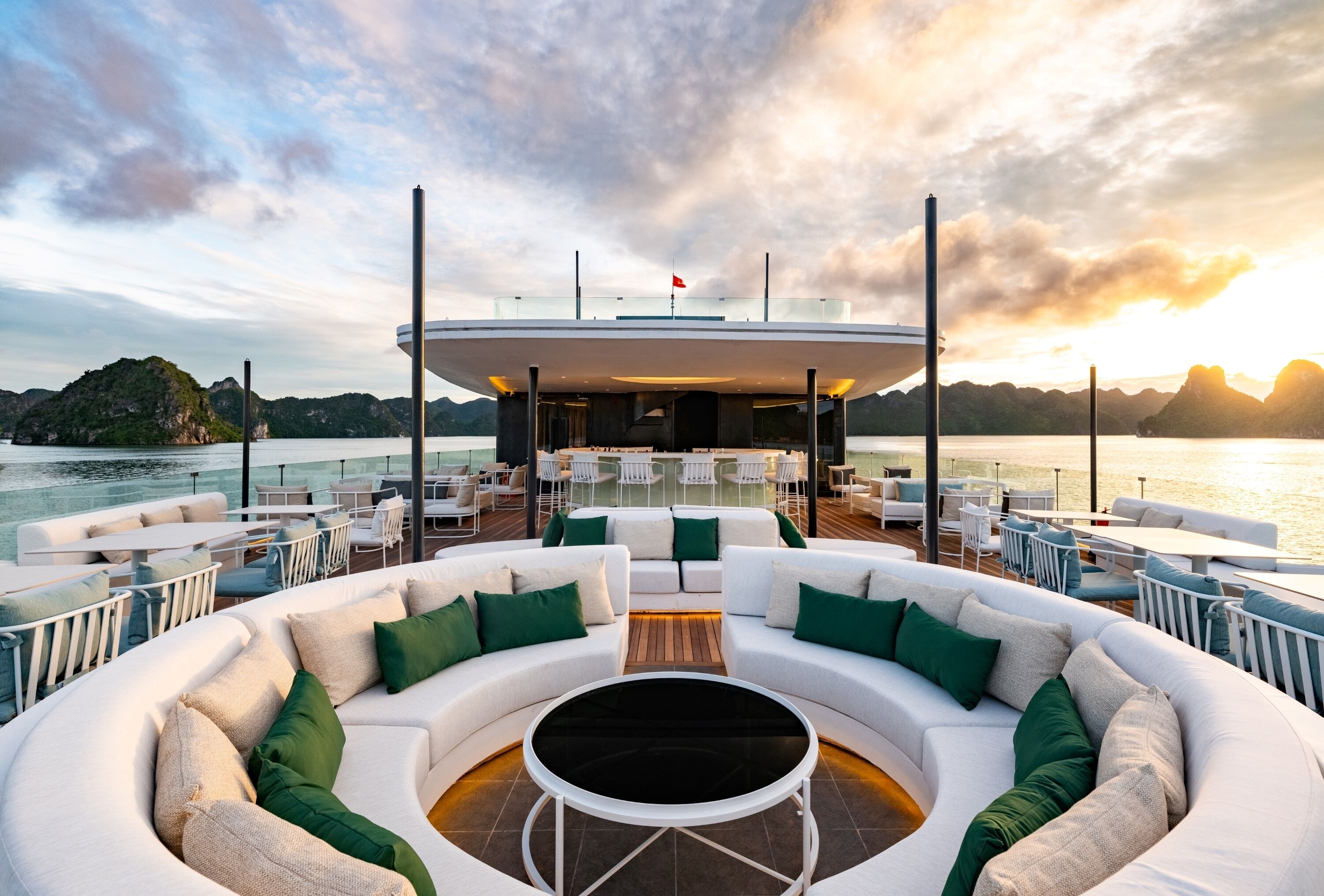 A modern outdoor lounge area with a circular seating arrangement, a central coffee table, and a view of the sea and mountains in the background.