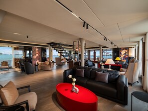 Un área de sala amplia con diseño moderno, que incluye una mesa de centro roja, asientos cómodos y grandes ventanales con vistas al mar.