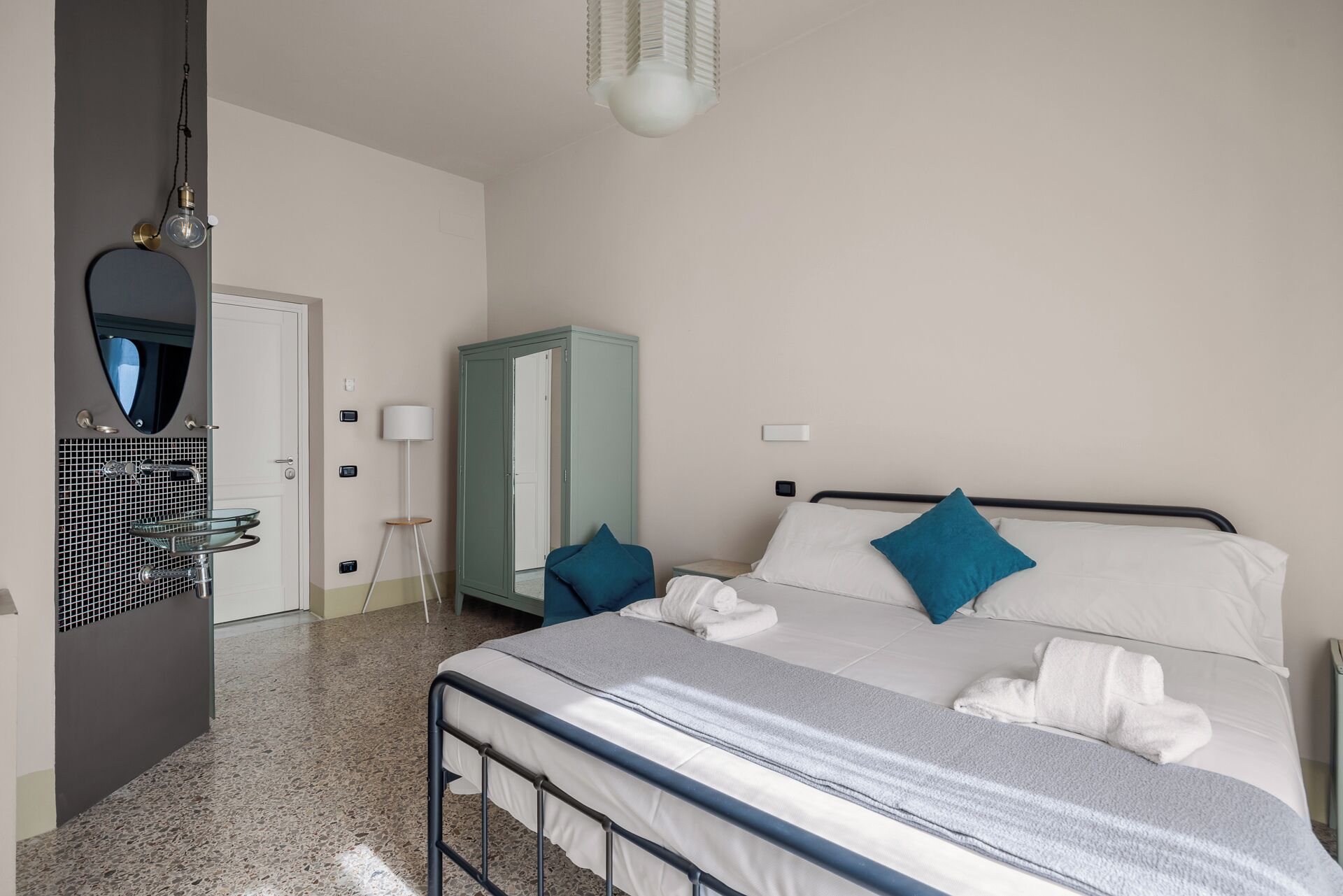 A hotel room with a bed, a wardrobe, a mirror, a sink, and a lamp.