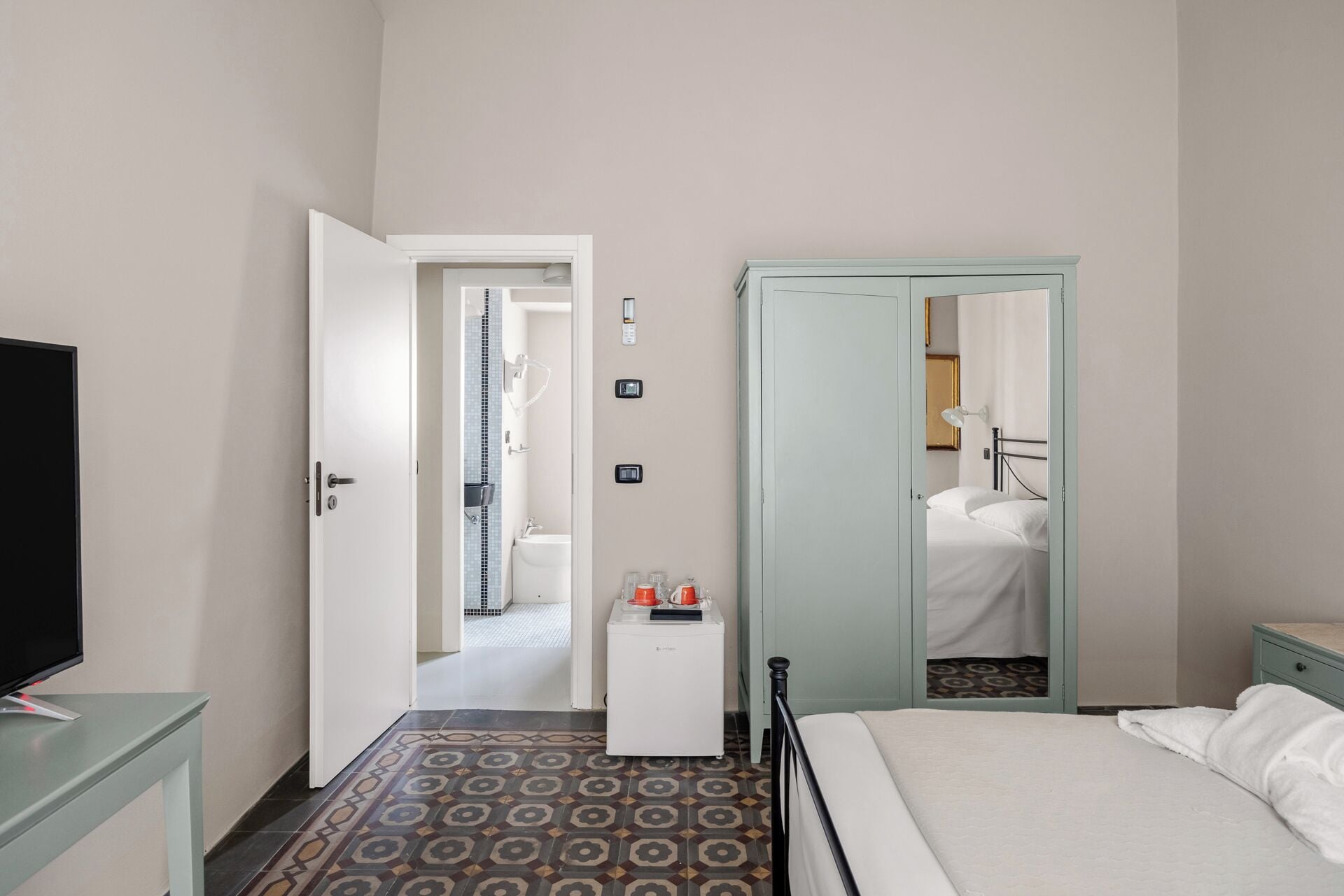 Habitación de hotel con una cama, un televisor, una mesita con una tetera, un espejo y una puerta que da a un baño.