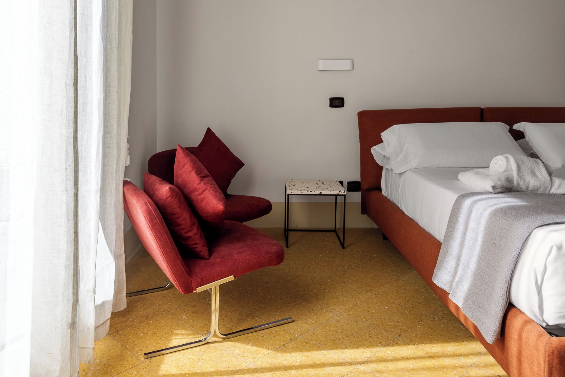 Ein Hotelzimmer mit einem Bett, einem roten Sessel, einem kleinen Tisch und einem Vorhang.
