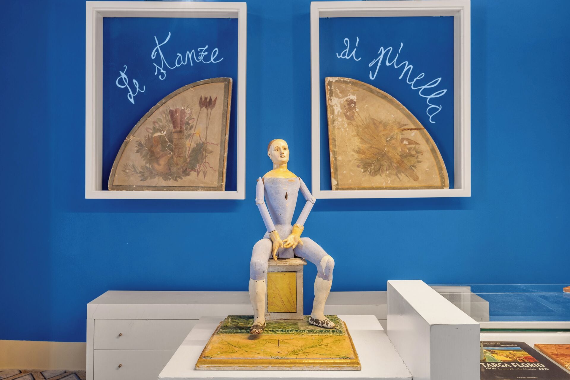 Eine Skulptur auf einem Sockel, mit einer blauen Wand und zwei gerahmten Kunstwerken darüber.
