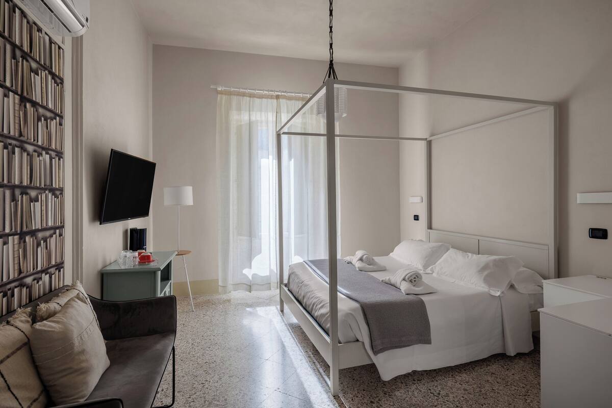 A modern hotel room with a four-poster bed, a flat-screen TV, a bookshelf, and a small table with a lamp.