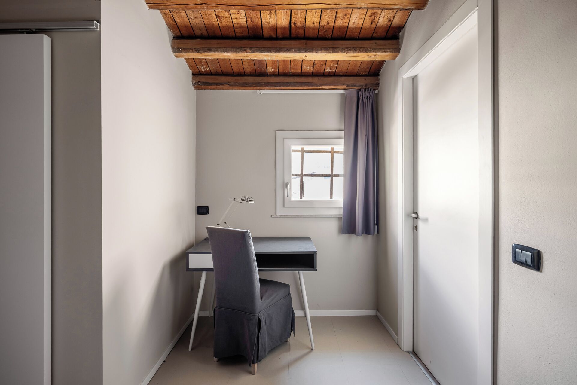 A small, narrow room with a wooden ceiling, a desk with a chair, a window with a curtain, and a door.