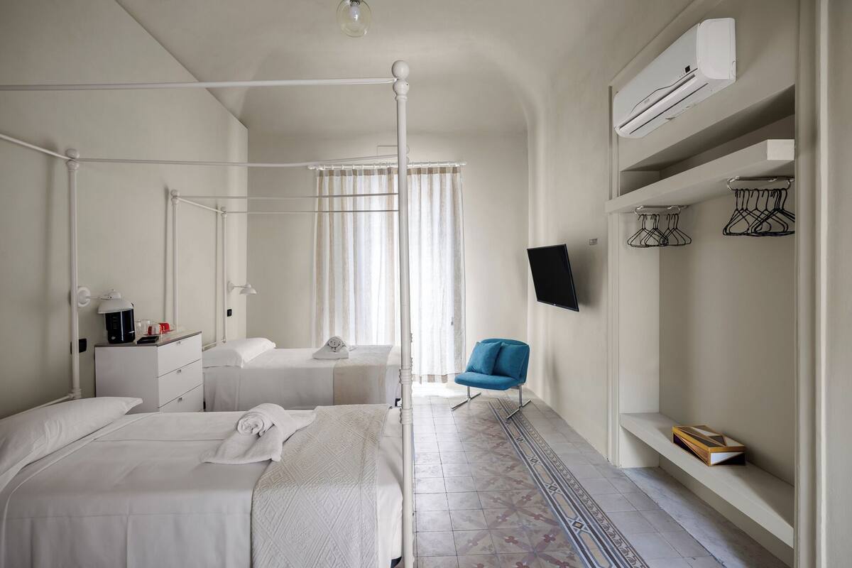 A hotel room with a bed, bedside table, lamp, air conditioning unit, TV, and a chair.