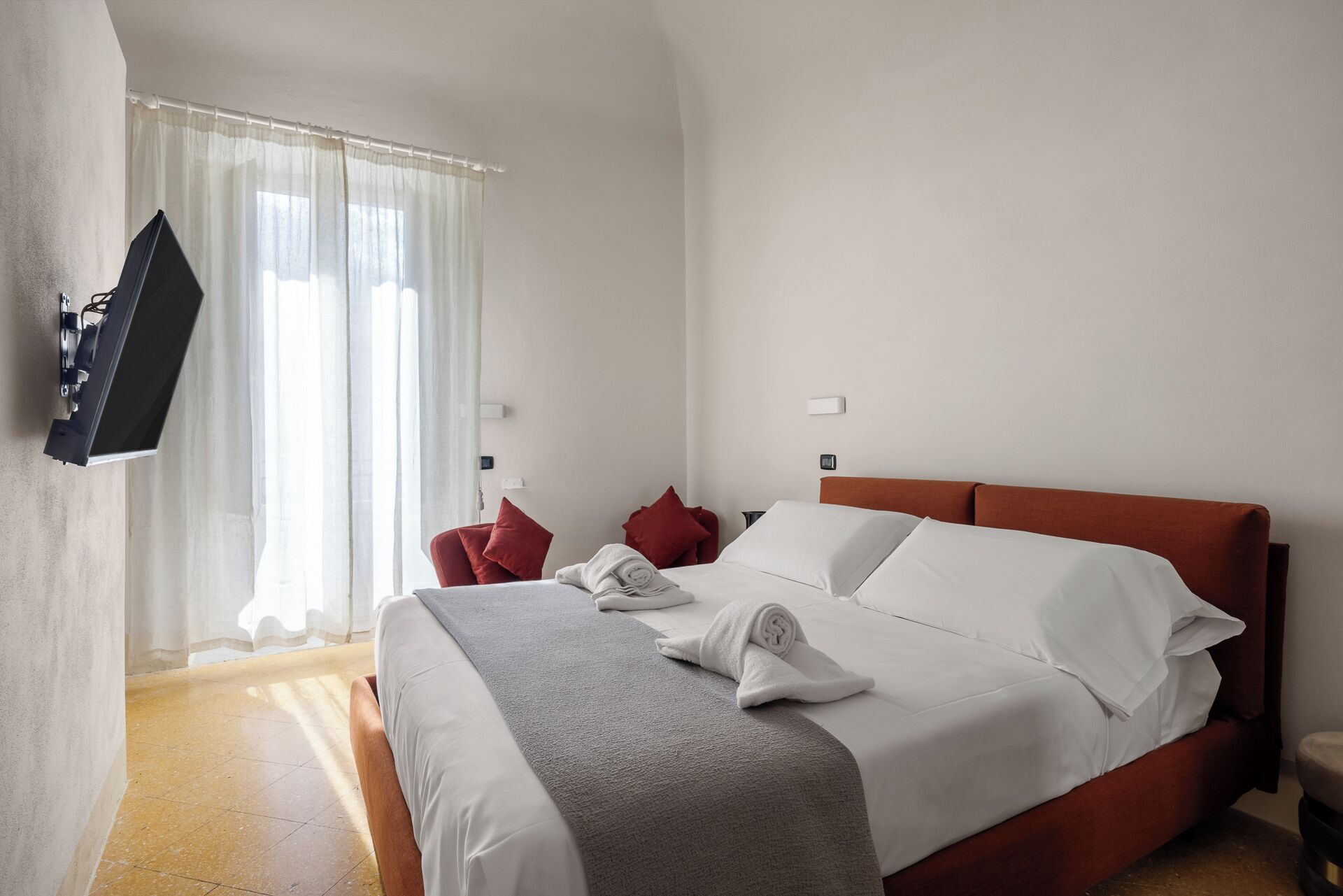 Quarto de hotel com cama, televisão fixada na parede e janela com cortinas.