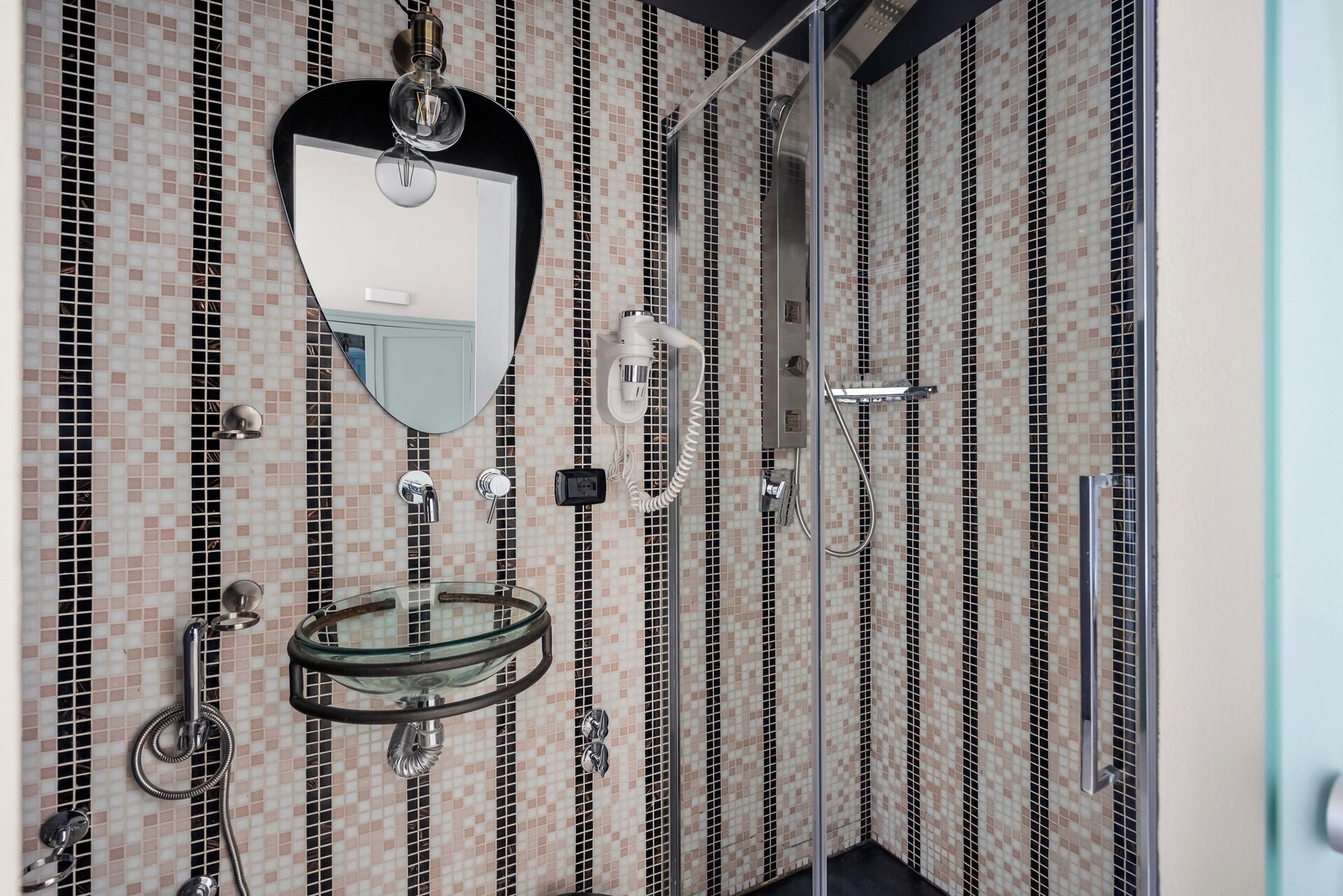 A bathroom with a glass sink, a mirror, a hairdryer, and a shower with a tiled wall.