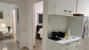 Quarto de hotel moderno com minibar branco, uma cama, escrivaninha com luminária e um quadro na parede.