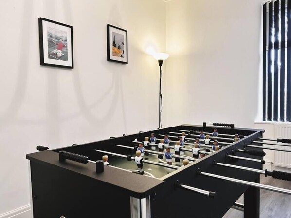 A foosball table in a room with white walls, two framed pictures, and a standing lamp.