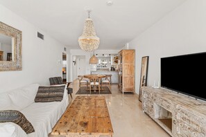 A living room with a sofa, coffee table, TV, and a dining area with chairs and a chandelier.