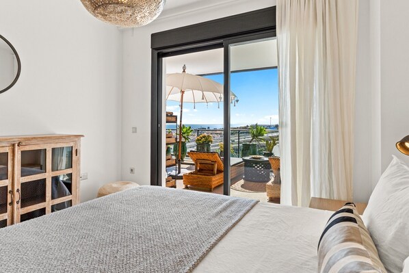 A bedroom with a large bed, a wooden cabinet, and a balcony with a view of the sea.