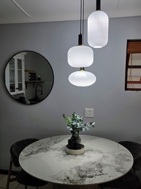A round dining table with a vase of green plants, three pendant lights, and two chairs.
