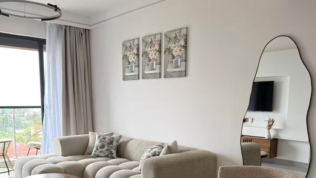 Ein modernes Wohnzimmer mit einem Ecksofa, einem runden Couchtisch und einem Esstisch mit Stühlen. An der Wand hängen drei gerahmte Kunstwerke, ein großer Spiegel und ein Flachbildfernseher.