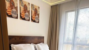 Ein ordentlich bezogenes Bett mit weißen Leinen und einer beigen Tagesdecke, ein Holz-Kopfbrett und ein großes Fenster mit Vorhängen.