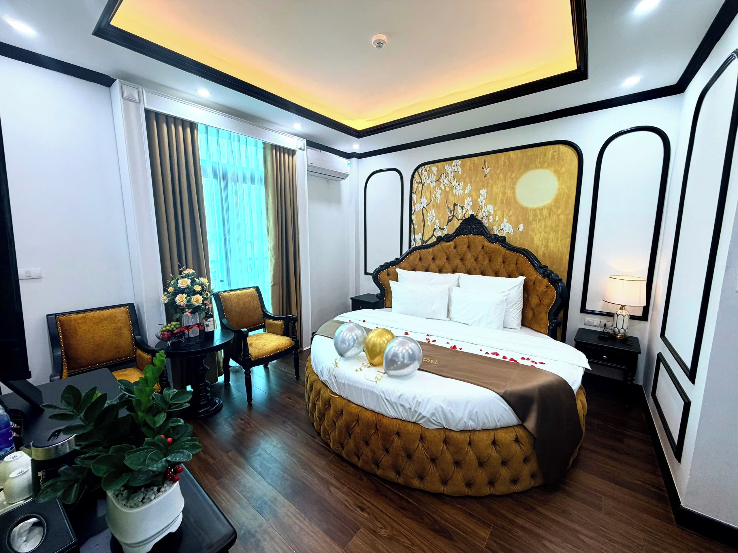 A hotel room with a large bed, a desk with a chair, a potted plant, and a window with curtains.